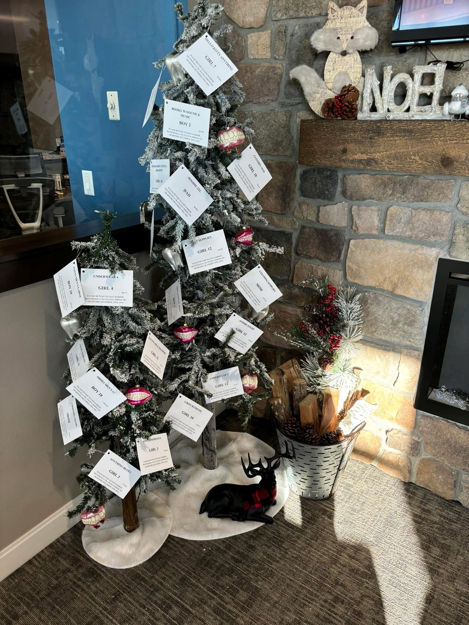 Two small artificial Christmas trees decorated with white cards detailing gift requests, next to a black deer figurine with a red plaid scarf and a metal bucket holding firewood and pine branches by a stone fireplace.
