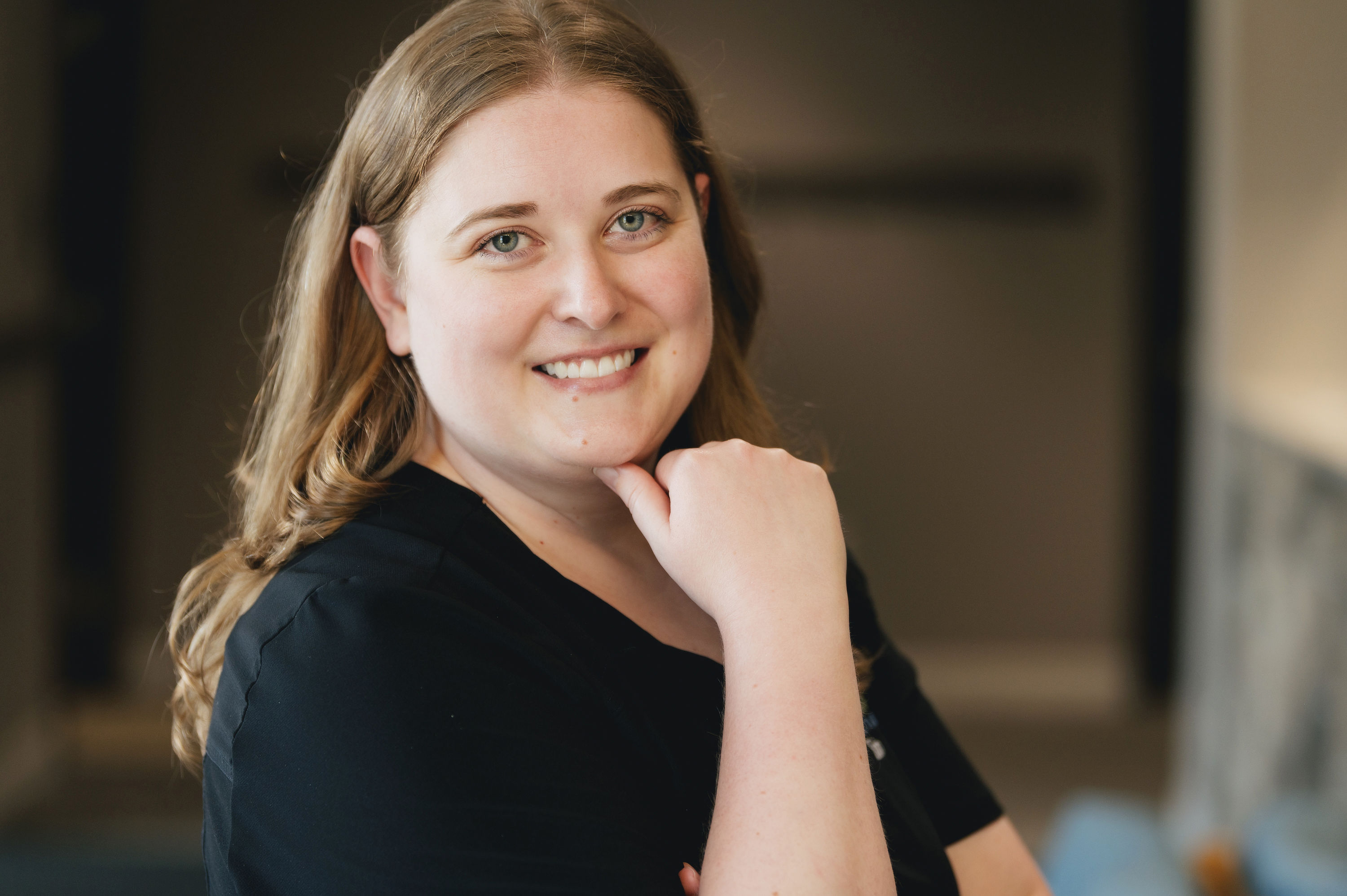 Smiling woman with wavy light brown hair resting her chin on her hand against a blurred indoor background.