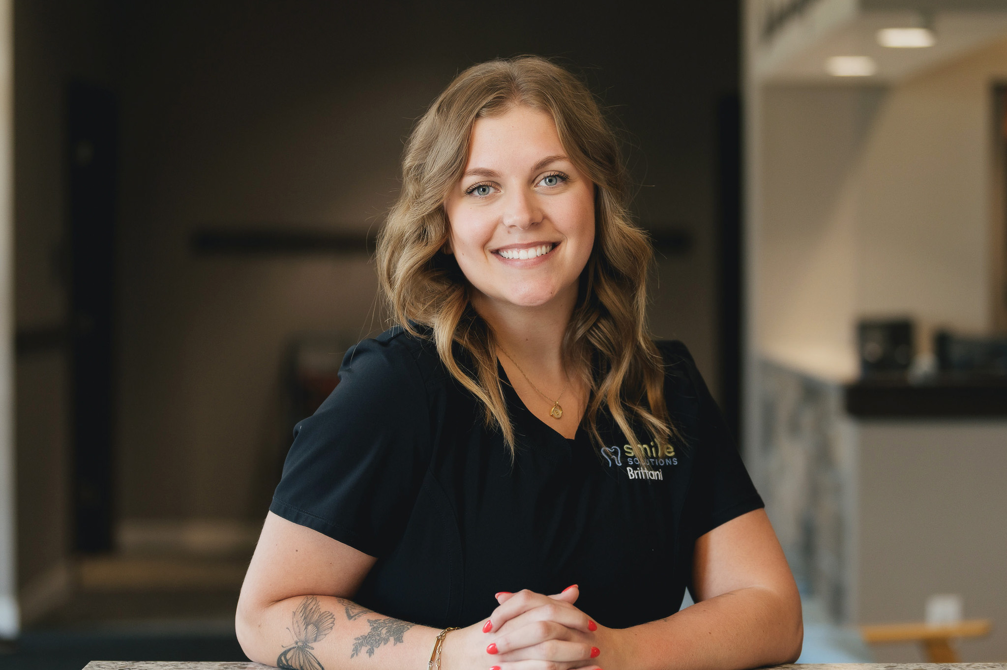 Smiling woman with wavy blonde hair, wearing a black shirt with 'Smile Solutions Brittani' logo, sitting at a table with tattooed forearms and clasped hands.
