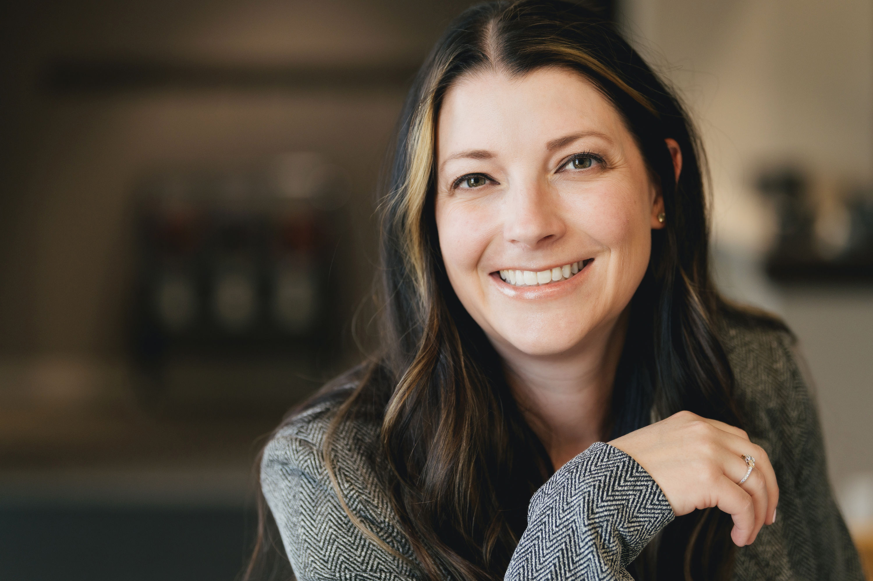 Smiling woman with long dark hair and light highlights, wearing a patterned jacket and a ring on her finger.