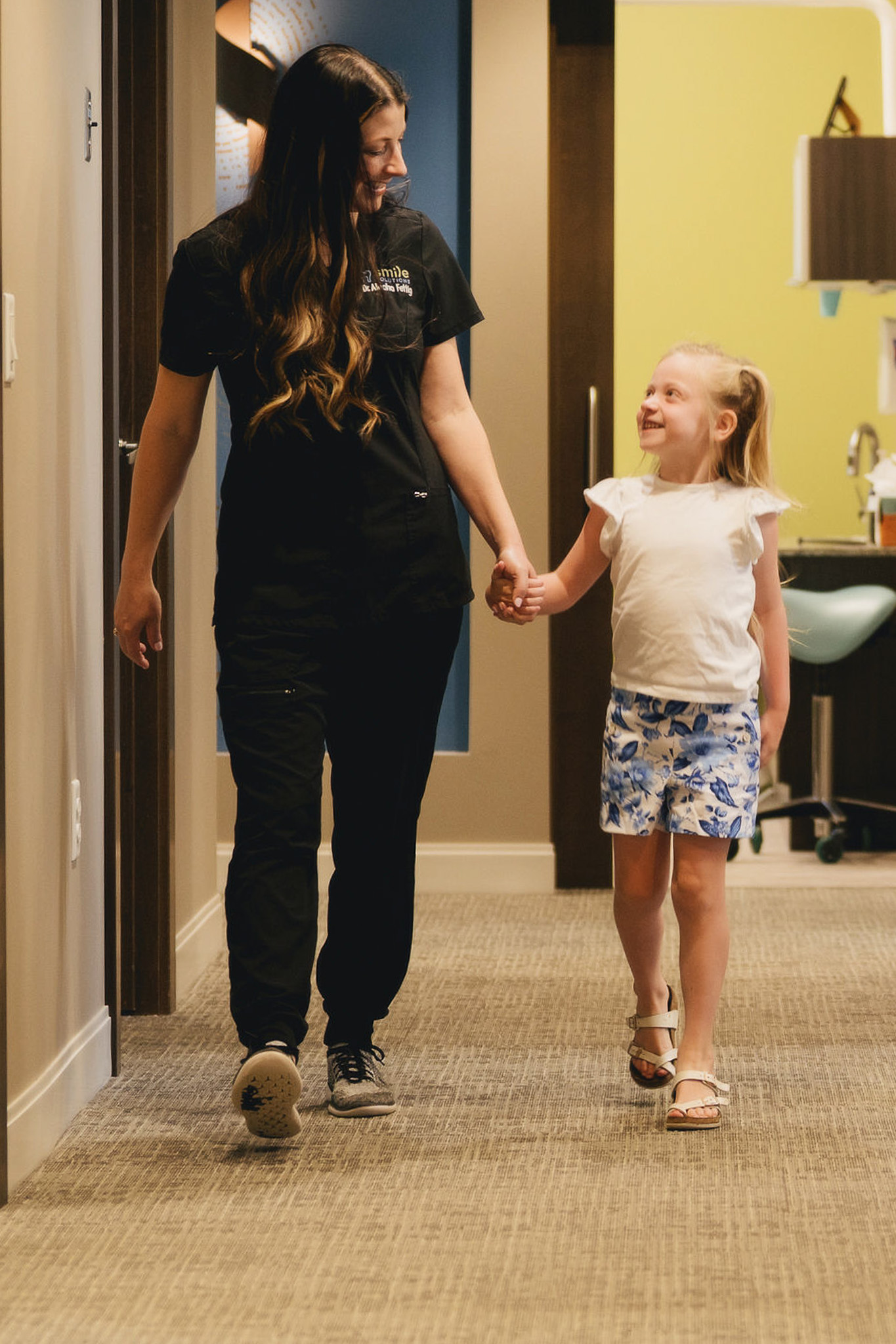 Woman in black scrubs holding hands and smiling at a young girl in a white top and floral shorts while walking down a hallway.