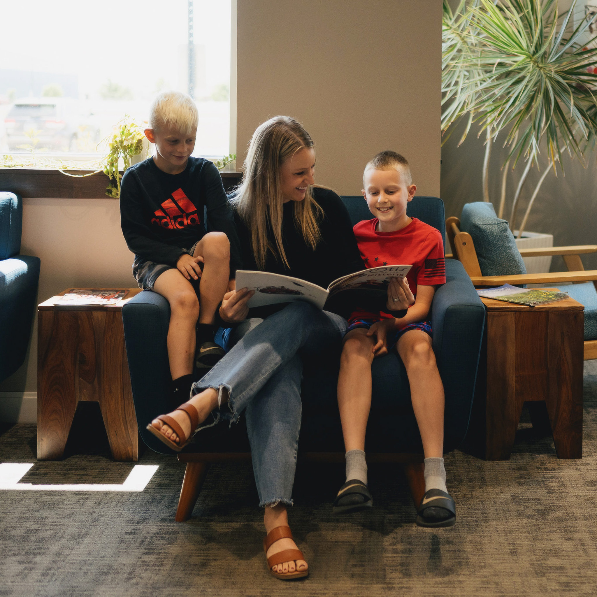 Woman sitting on a blue couch reading a book with two smiling boys, one on each side.