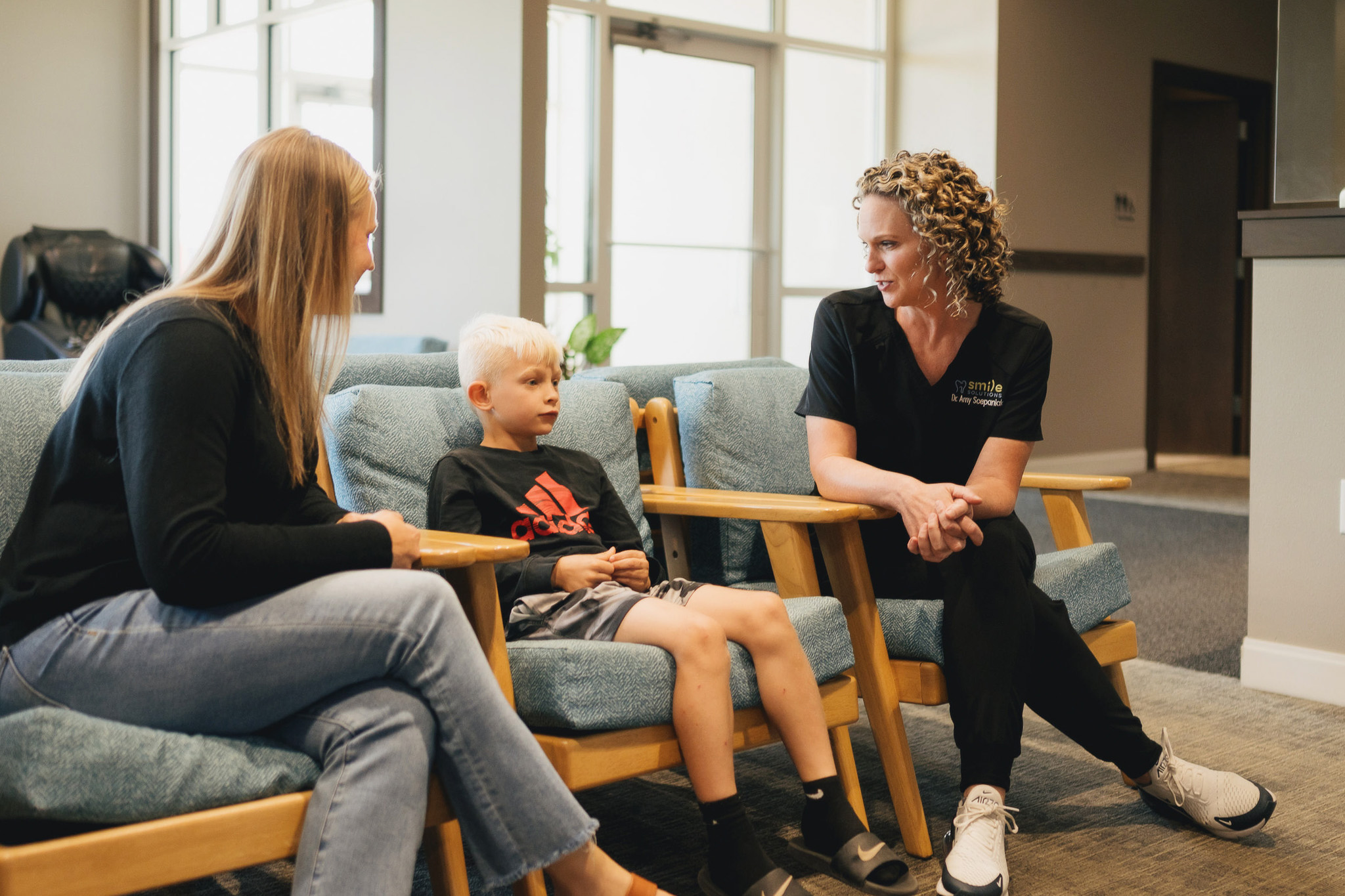 A woman and a boy sitting on chairs in a waiting area talking to a female healthcare professional wearing scrubs.