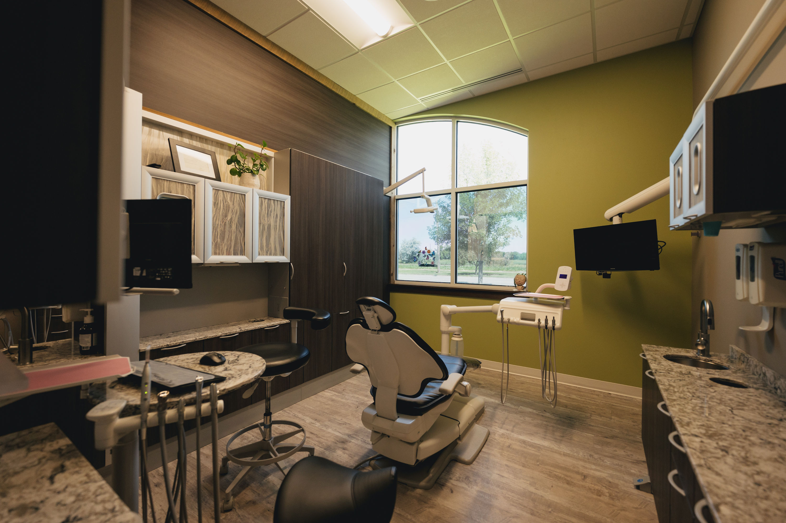Modern dental examination room with dental chair, equipment, and large window with a view of trees outside.
