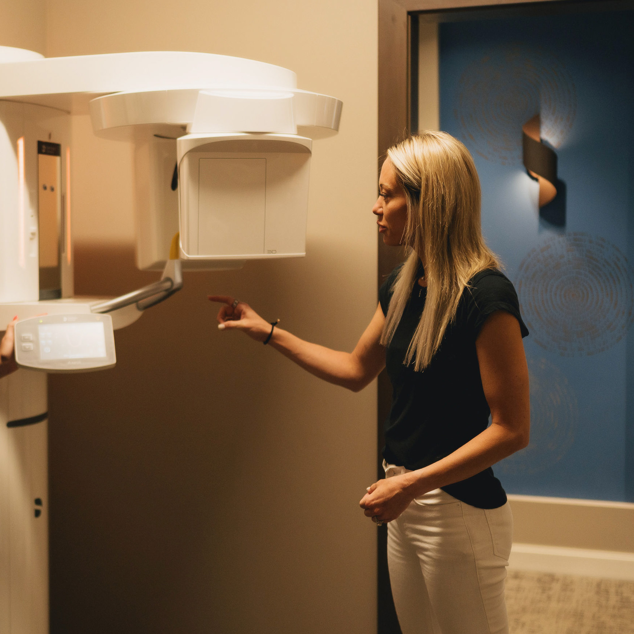Woman with long blonde hair pointing at controls of a large dental X-ray machine in a clinic room.