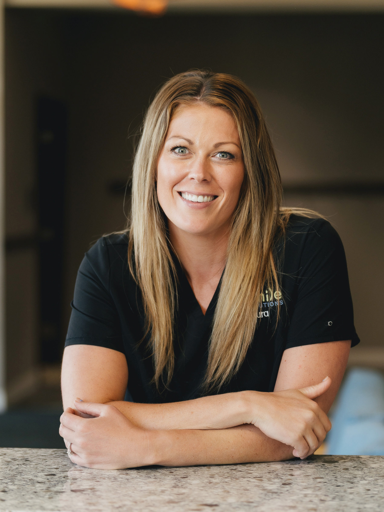 Woman with long blonde hair smiling and resting her crossed arms on a granite countertop, wearing a black shirt.