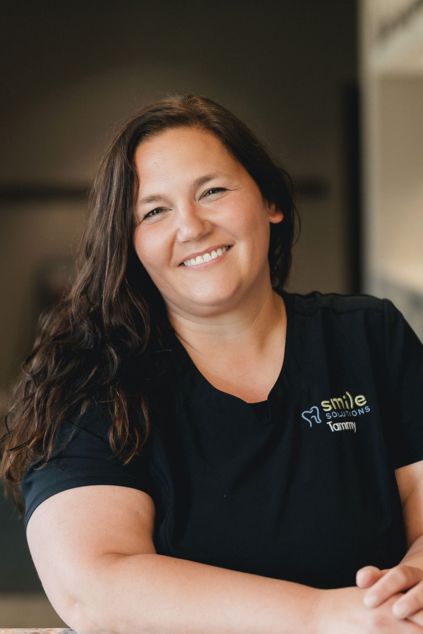 Smiling woman with long brown hair wearing a black Smile Solutions shirt with the name Tommy.
