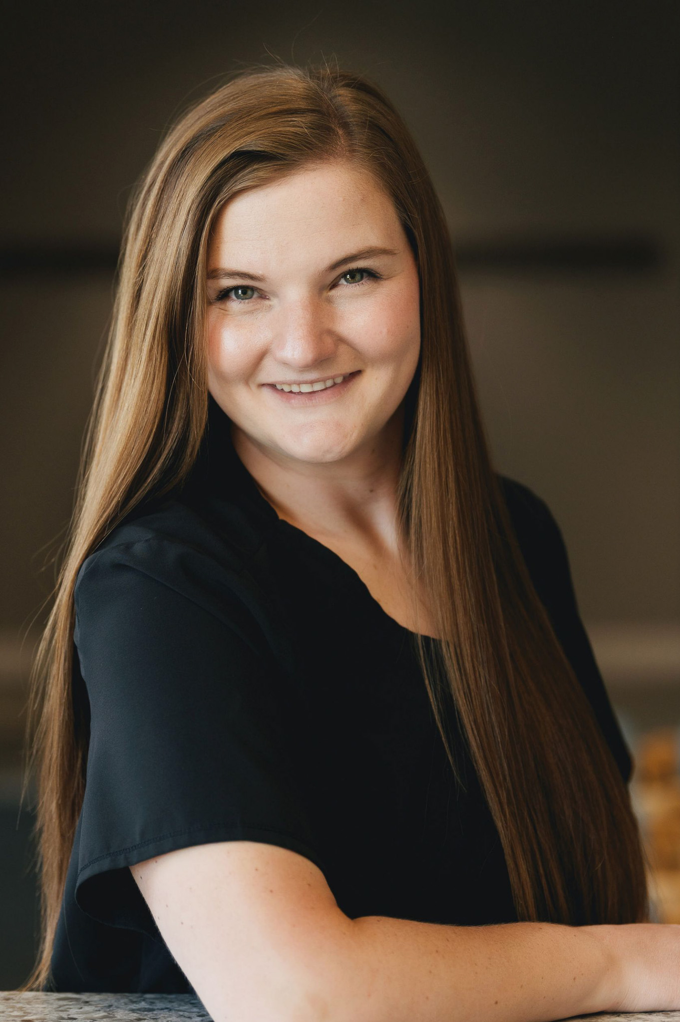 Smiling woman with long straight brown hair wearing a black top against a dark blurred background.
