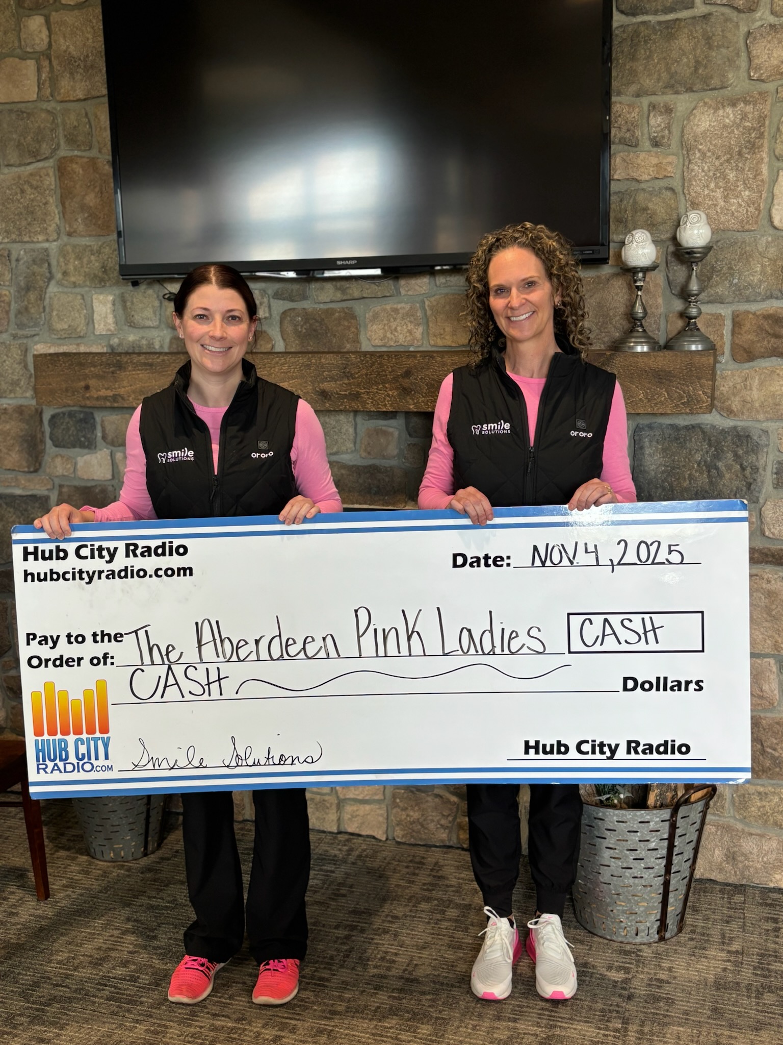 Two women wearing black vests and pink shirts holding a large check from Hub City Radio made out to The Aberdeen Pink Ladies dated November 4, 2025.