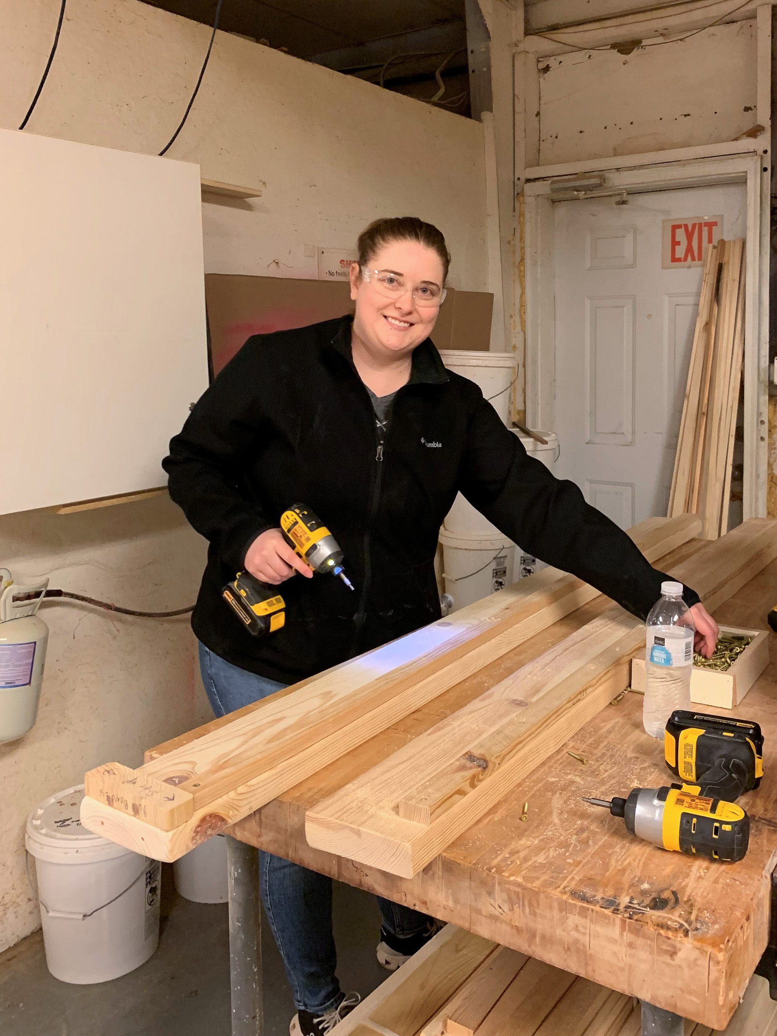 Woman wearing safety glasses and a black jacket holding a cordless drill while working with wooden planks on a workbench in a workshop.