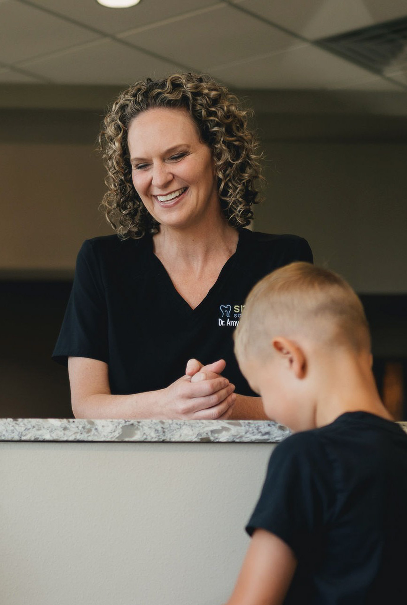 Smiling female dentist with curly hair wearing a black uniform stands behind a marble counter, interacting with a young boy facing away.