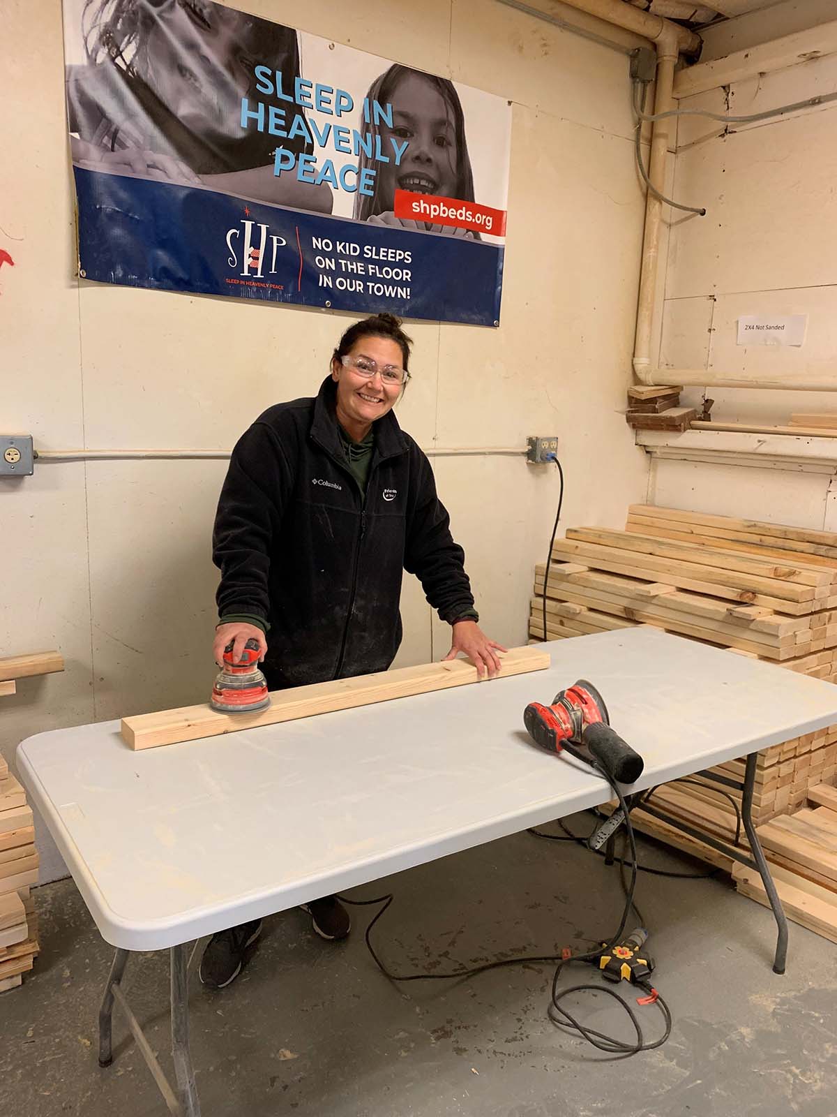 Woman wearing safety glasses sanding a wooden board on a table in a workshop with stacks of wood and a Sleep in Heavenly Peace banner on the wall.