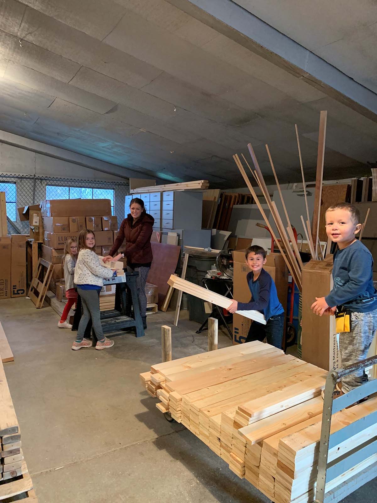 Four children and a woman handling wooden planks in a workshop with stacked lumber and cardboard boxes.