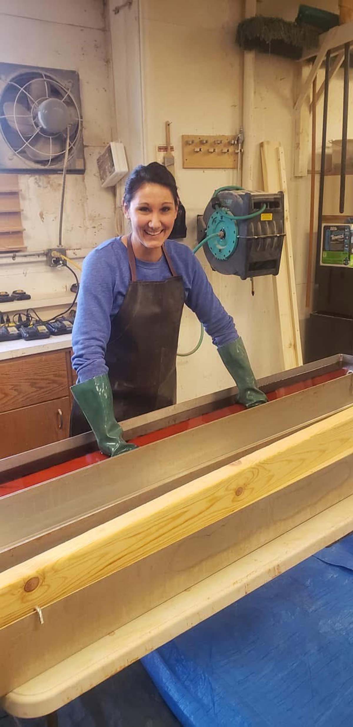 Woman wearing green gloves and a dark apron smiles while working at a long metal trough in a workshop.