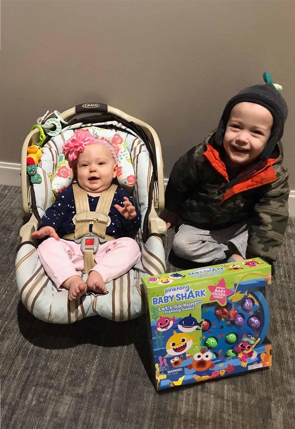 Baby in a car seat wearing a flower headband next to a smiling toddler in a camouflage jacket holding a Baby Shark fishing game box.