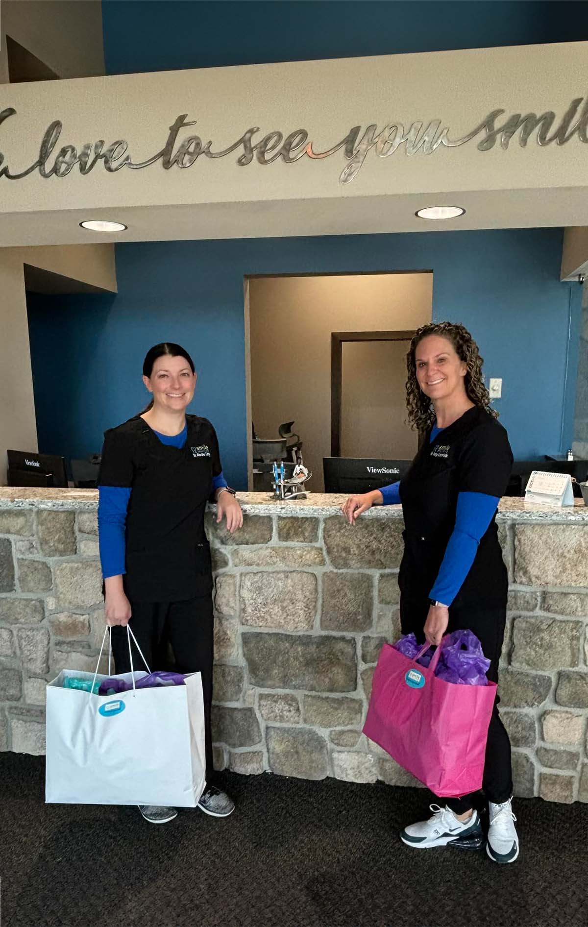 Two women wearing black scrubs and blue undershirts standing in front of a stone reception counter holding gift bags, smiling.