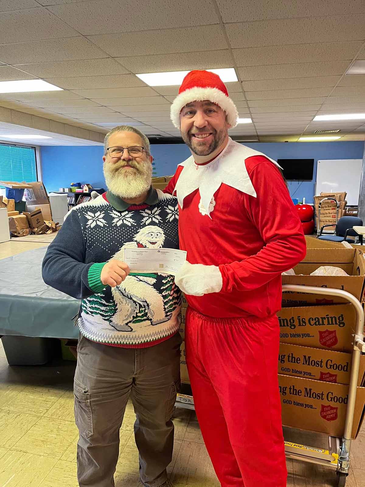 Two men standing indoors; one wearing a Christmas sweater with a Yeti design holding a check, the other dressed in a red elf costume with white gloves and hat.
