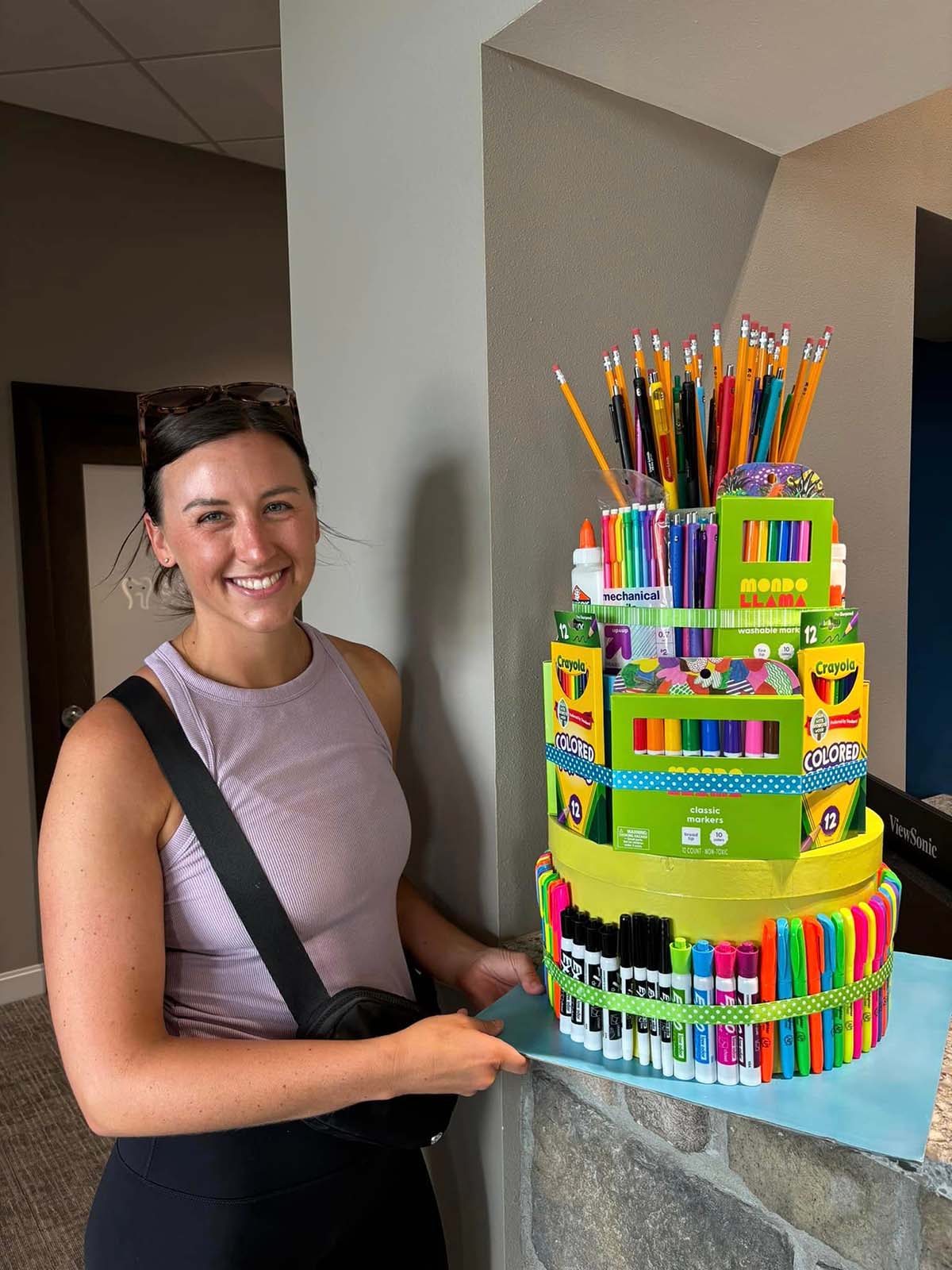 Smiling woman holding a colorful two-tiered art supply cake decorated with markers, colored pencils, and pencils.
