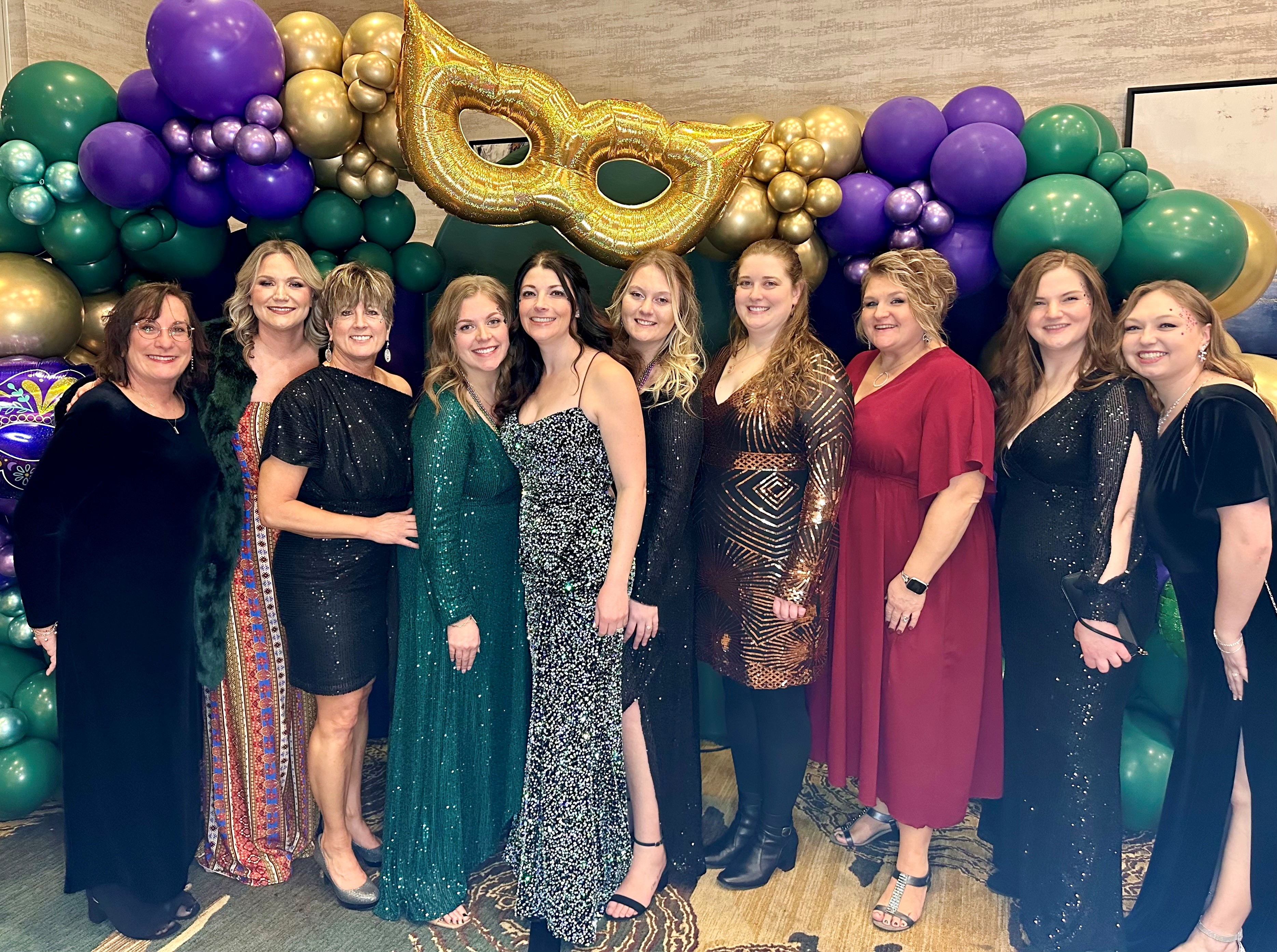 Ten women dressed in formal evening gowns posing in front of a colorful balloon arch with a large gold mask decoration.