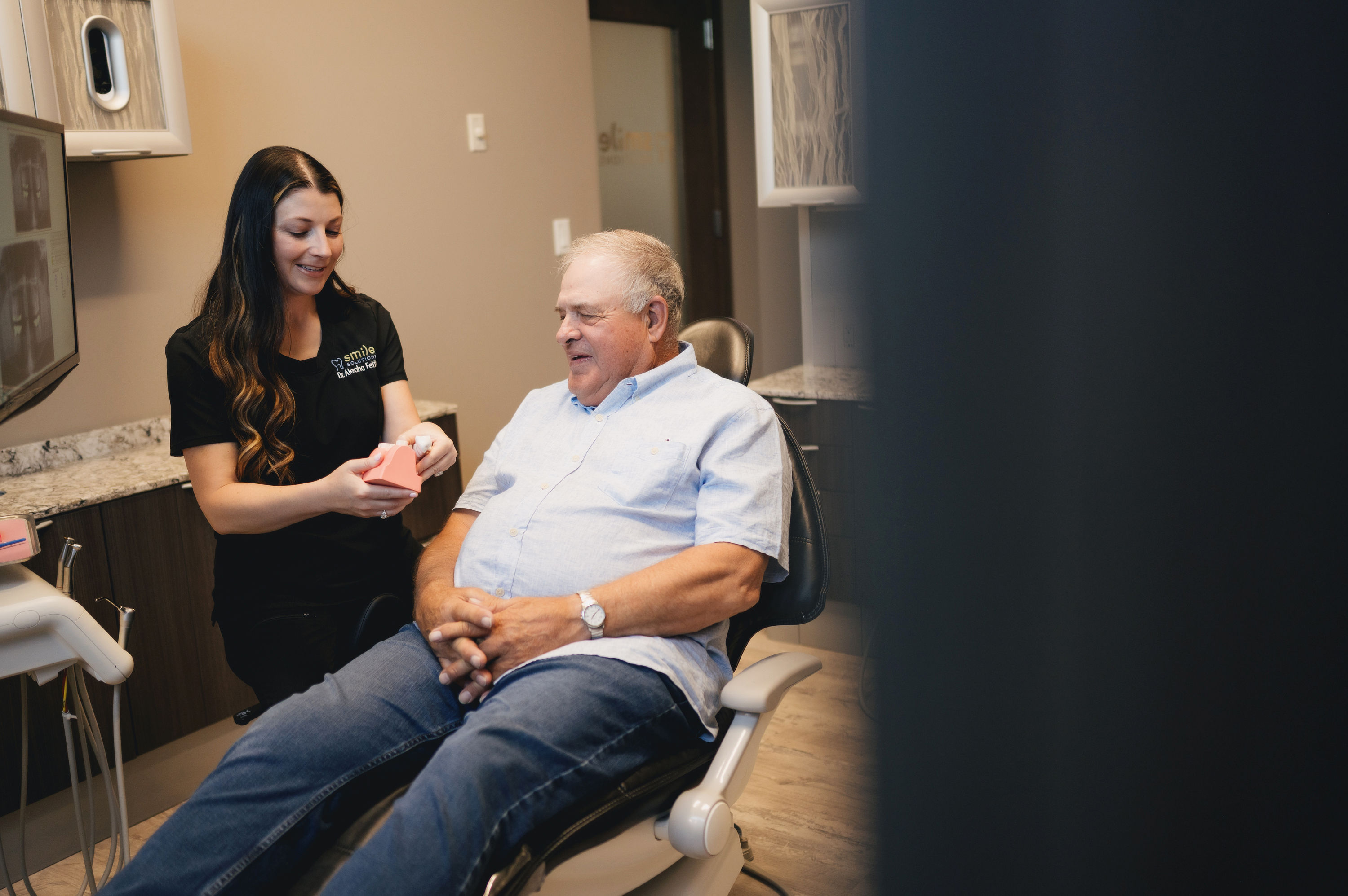 Female dentist explaining dental model to an elderly male patient seated in a dental chair.
