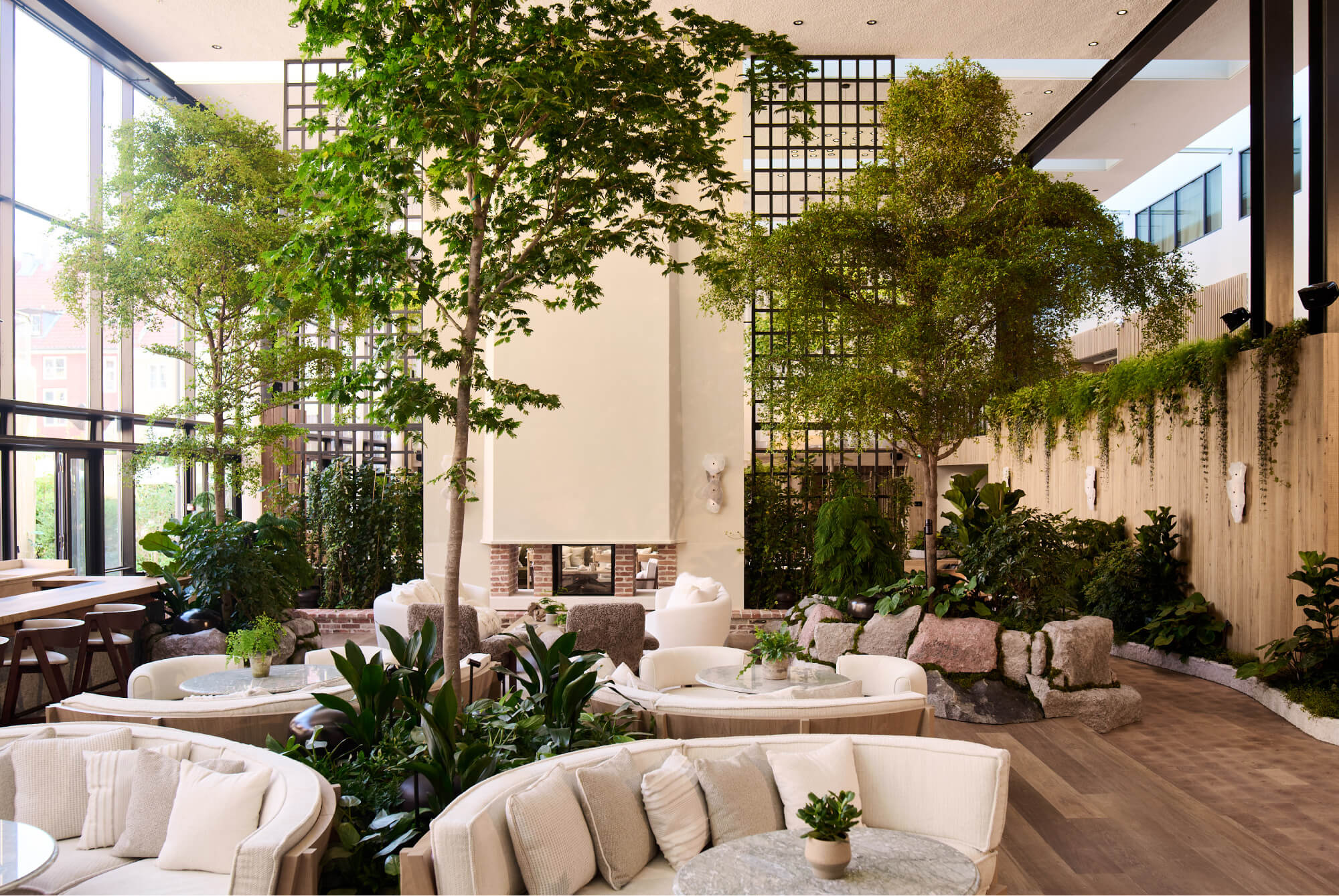 Indoor lounge area with round cream sofas, plants, trees, and a modern fireplace on a wooden floor.