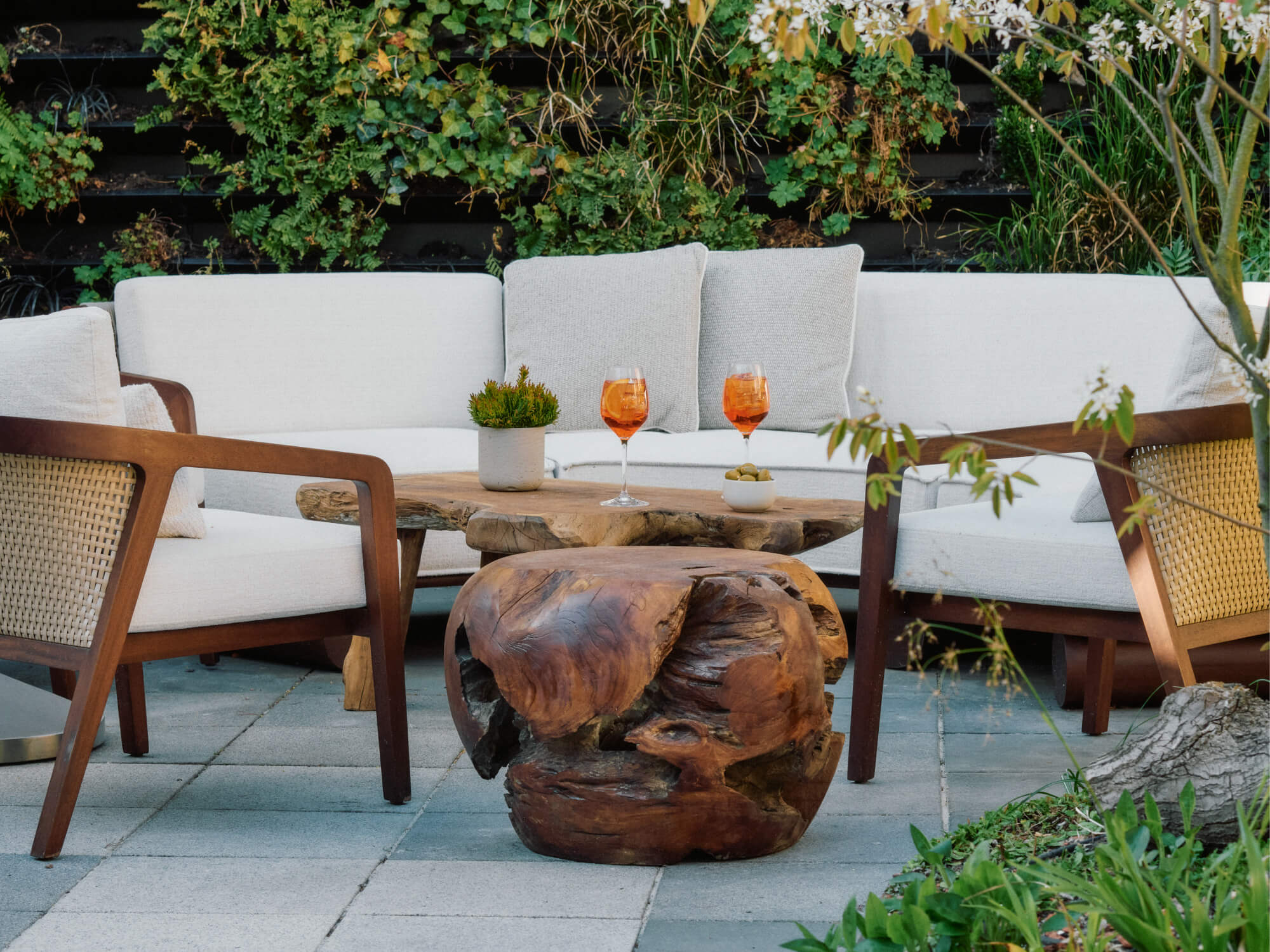 Outdoor seating area with wooden chairs, a sofa, rustic wood tables, drinks, and green plants.