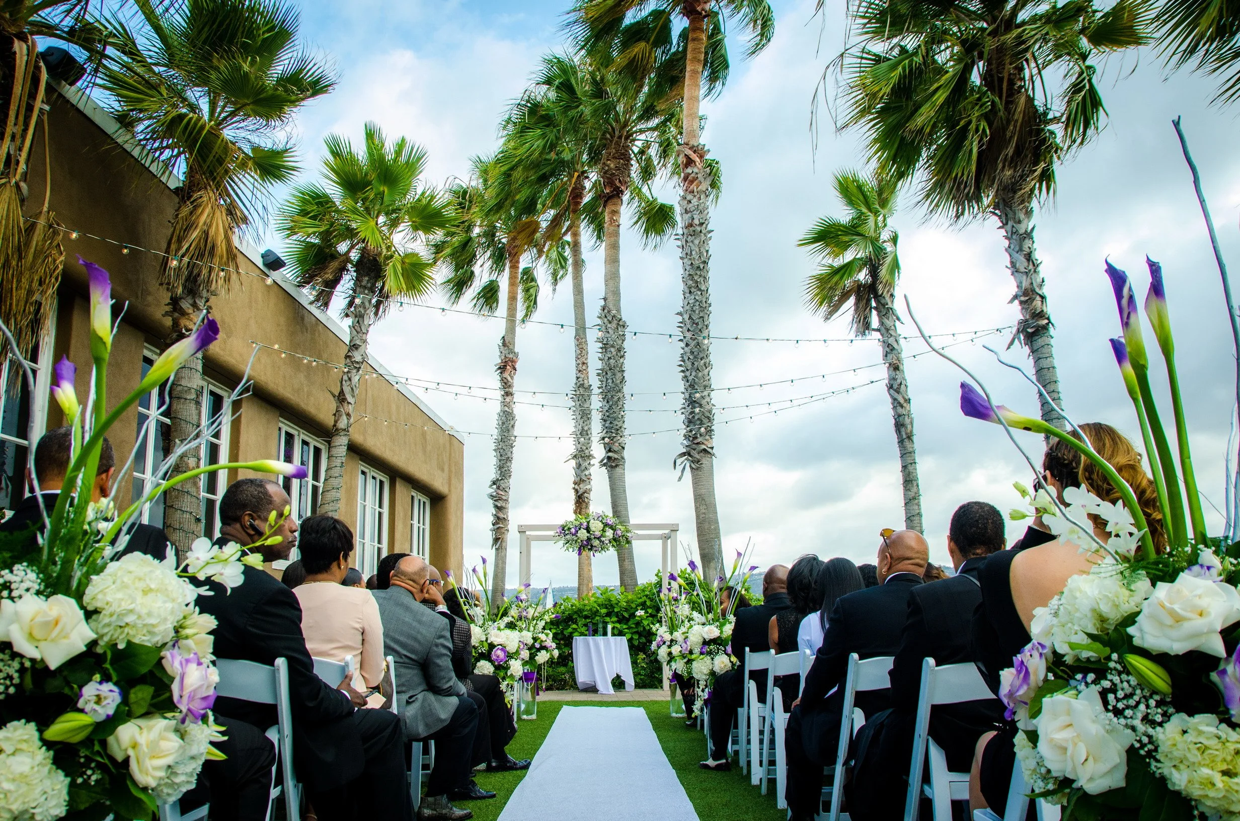 Outdoor wedding ceremony setup with guests seated on white chairs along an aisle lined with white and purple floral arrangements