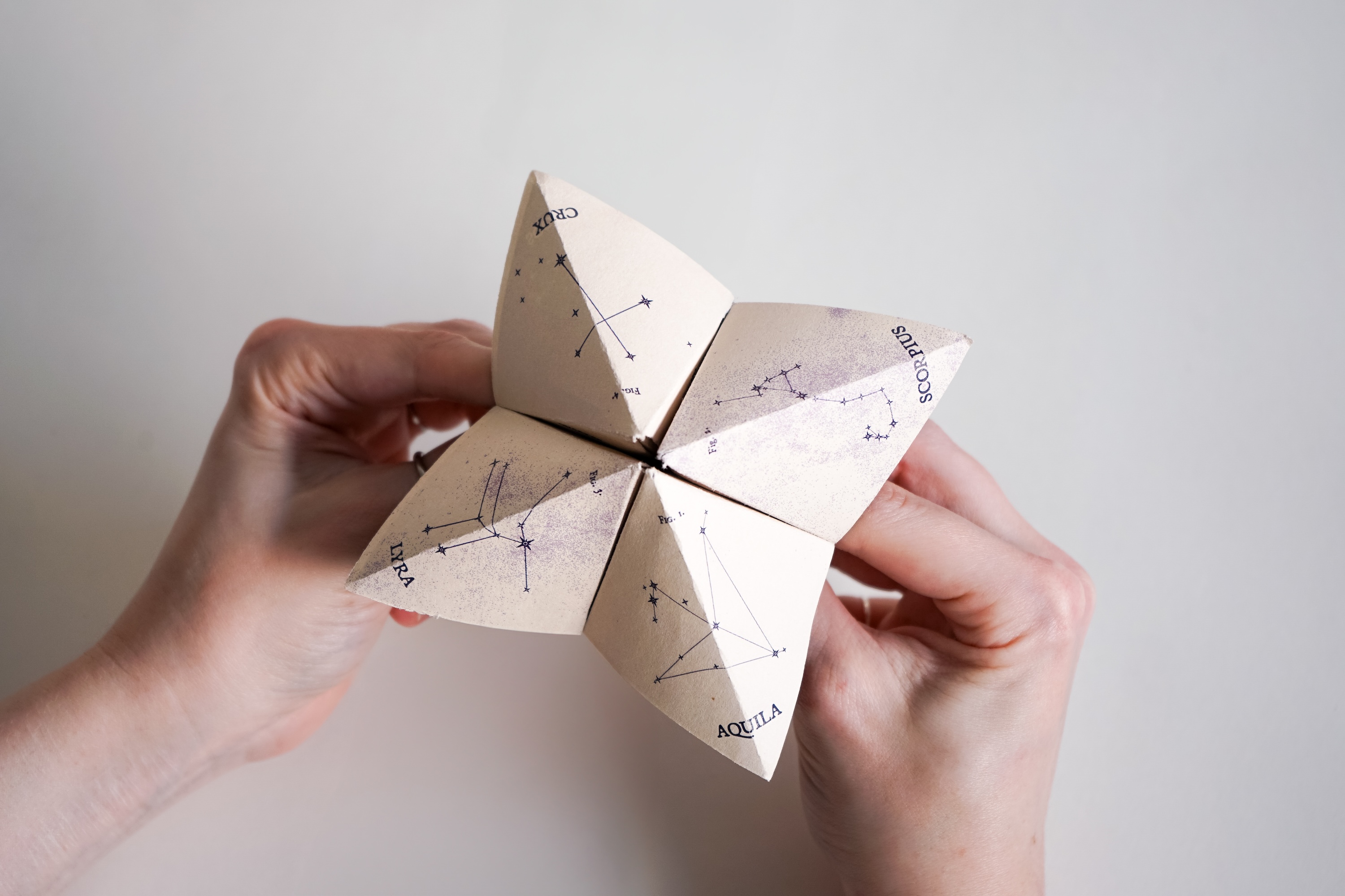 Hands holding a paper fortune teller with illustrations of star constellations labeled Crux, Scorpius, Lyra, and Aquila.