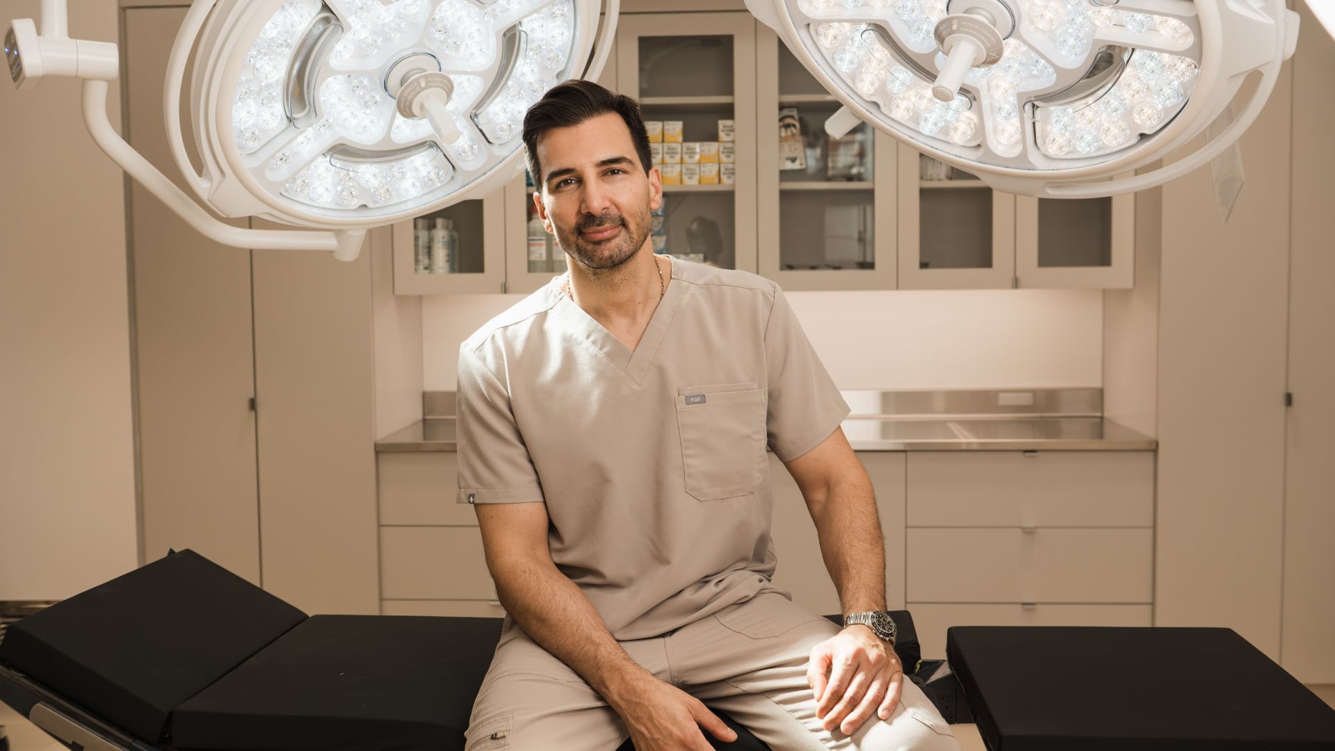 Dr. Motakis in scrubs sitting on an operating table in a bright surgical room.