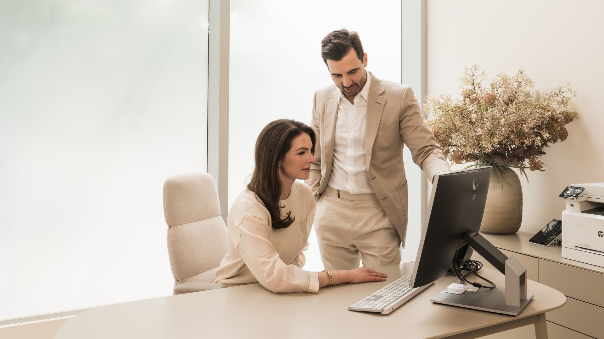 Dr. Motakis and Dr. Salsberg standing and woman in light sweater sitting and looking at computer monitor in modern office.