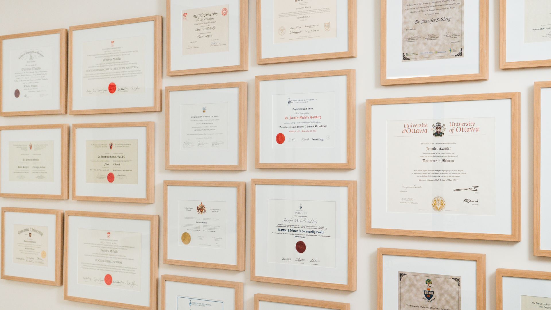 Wall displaying multiple framed medical and academic certificates and diplomas with wood-colored frames.