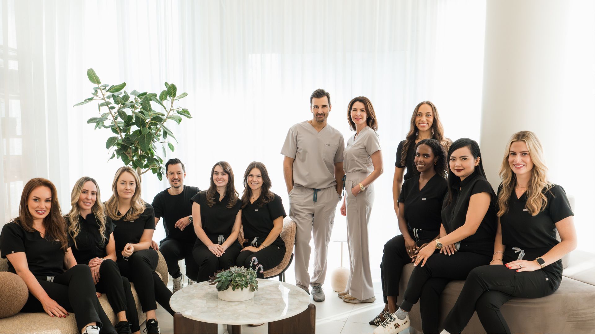 AvenueMD's team posing in a bright, modern office with white curtains, a plant, and a round table with a small succulent arrangement.
