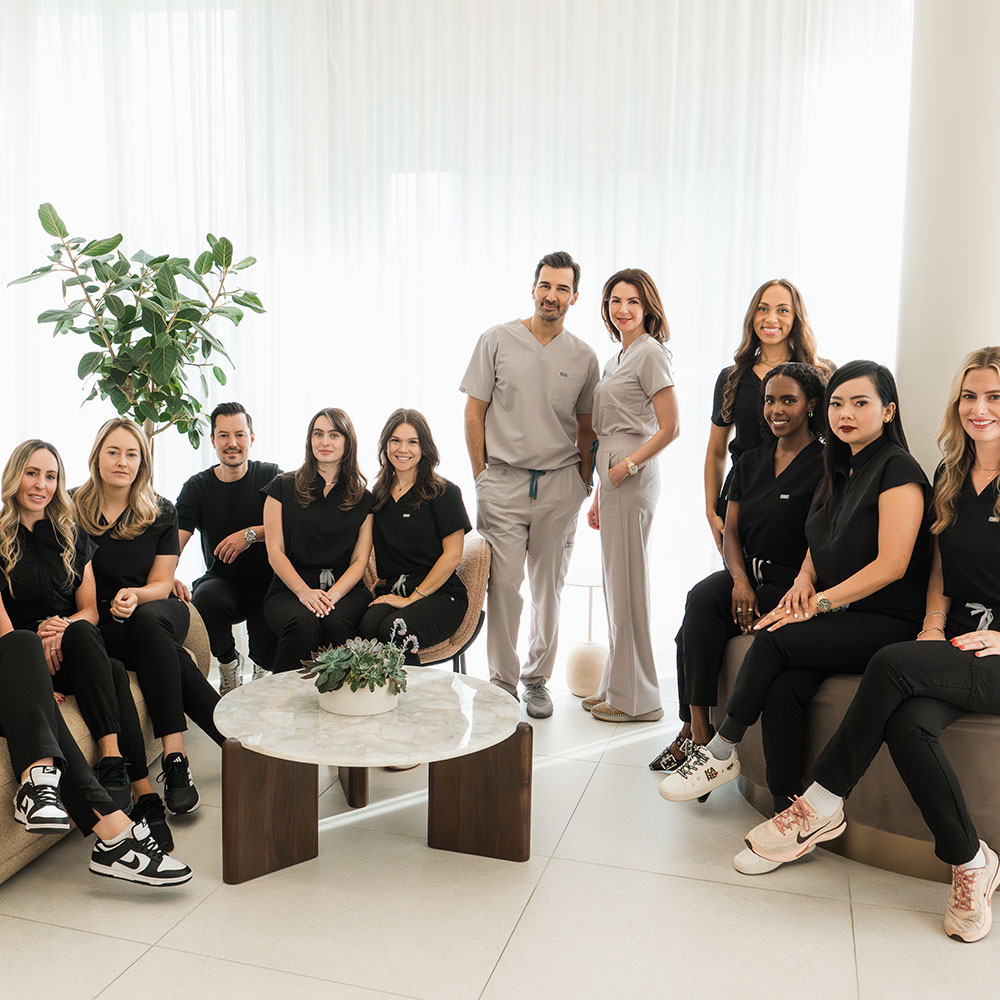AvenueMD's team posing in a bright, modern office with white curtains, a plant, and a round table with a small succulent arrangement.