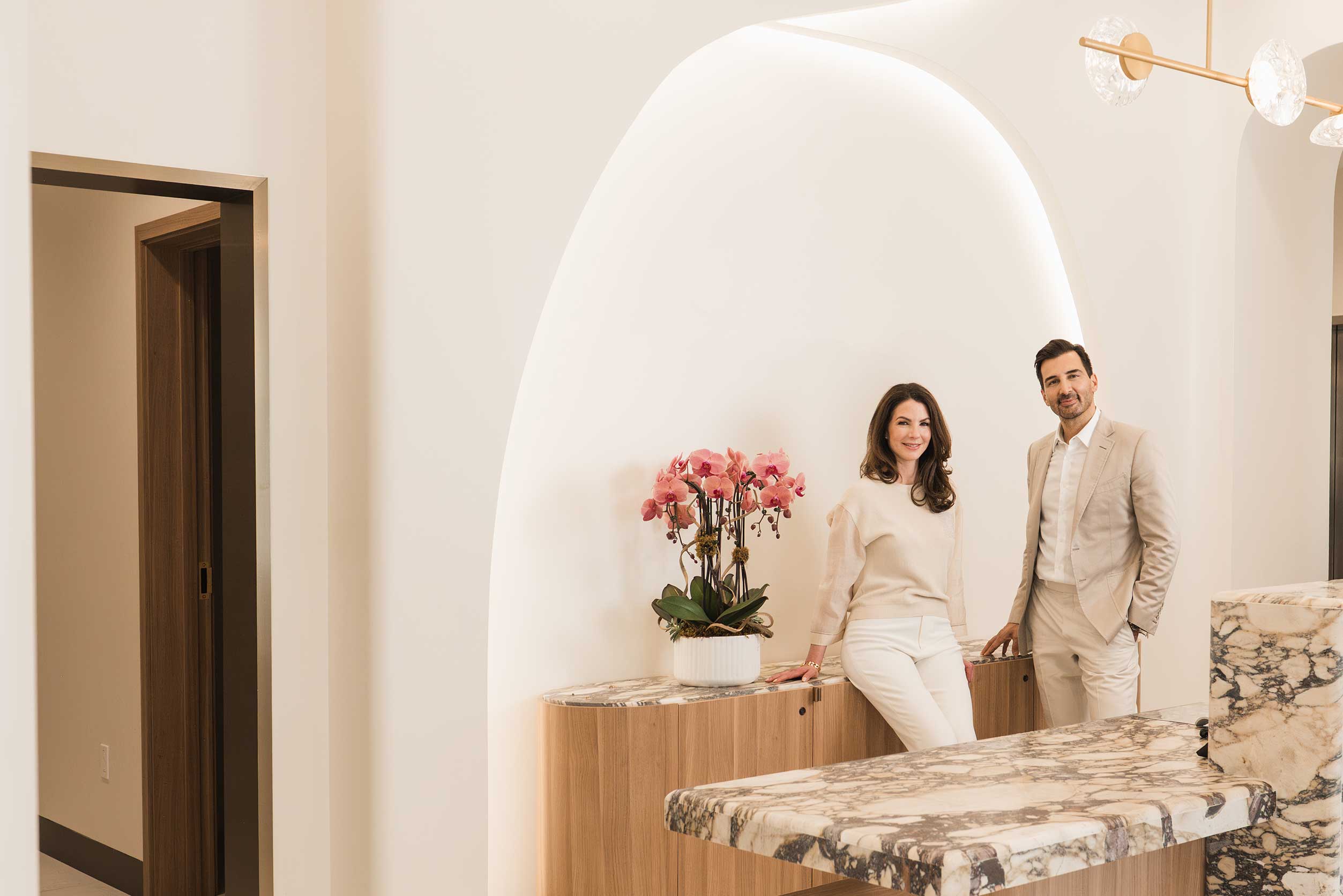 Dr. Motakis and Dr. Salsberg, both dressed in beige and white, standing and sitting respectively in a bright room with a marble countertop and a wooden cabinet with a vase of pink orchids.
