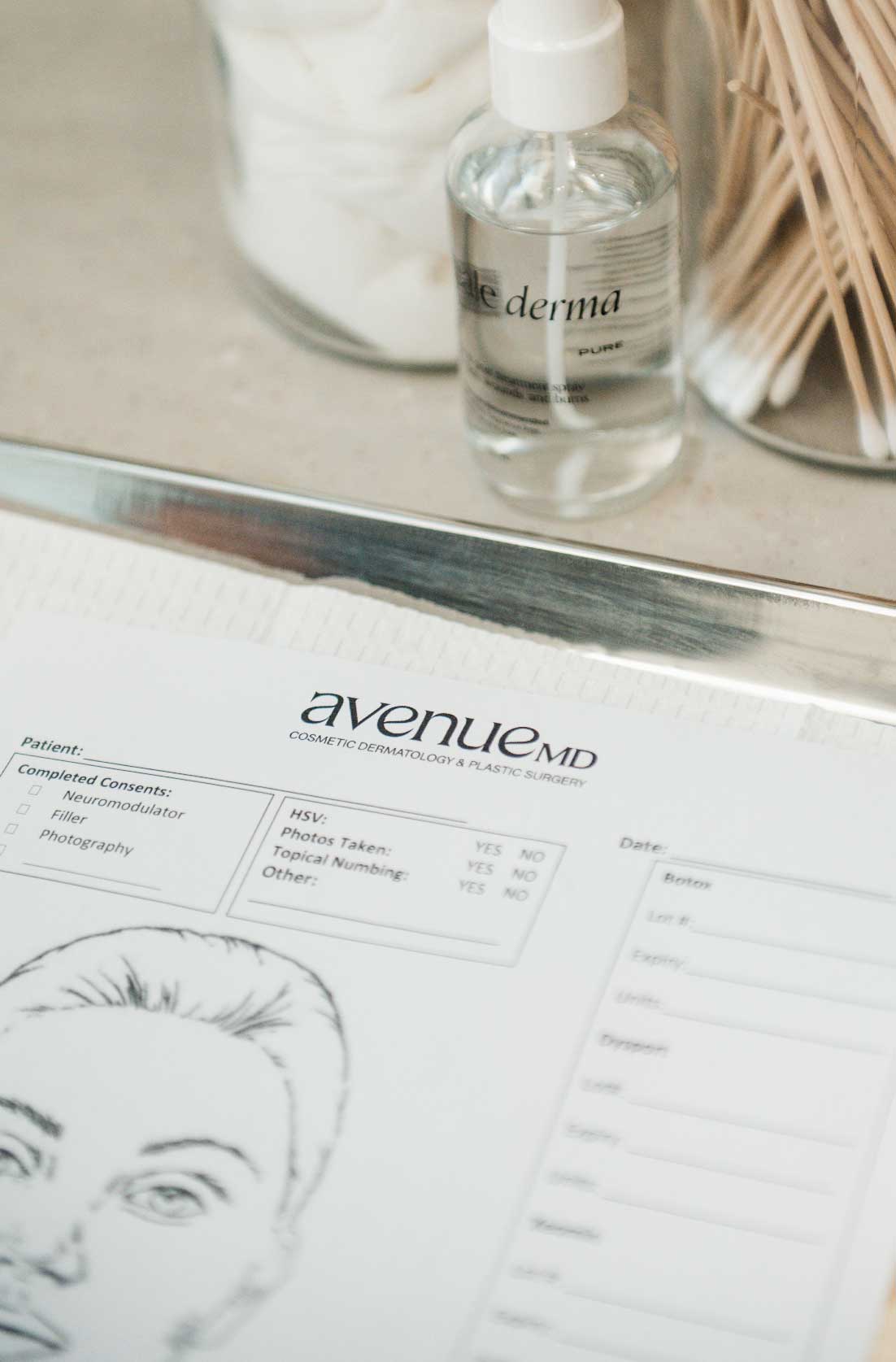 Close-up of a medical consent form from AvenueMD with a facial sketch, next to a clear bottle labeled 'derma' and a container of cotton swabs and pads.