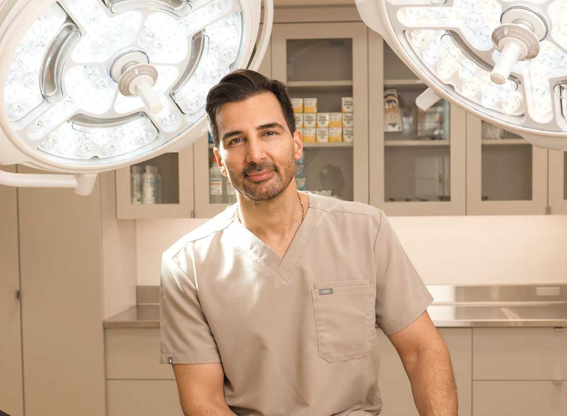 Dr. Motakis in beige scrubs sitting in a well-lit surgical room.