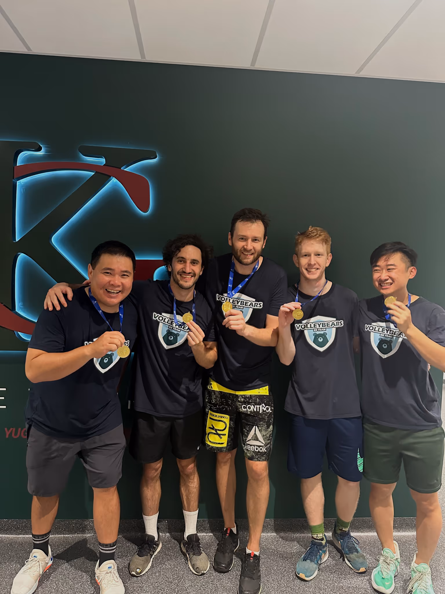 Five men standing in a row, smiling and holding gold medals, wearing Volleybears team shirts.