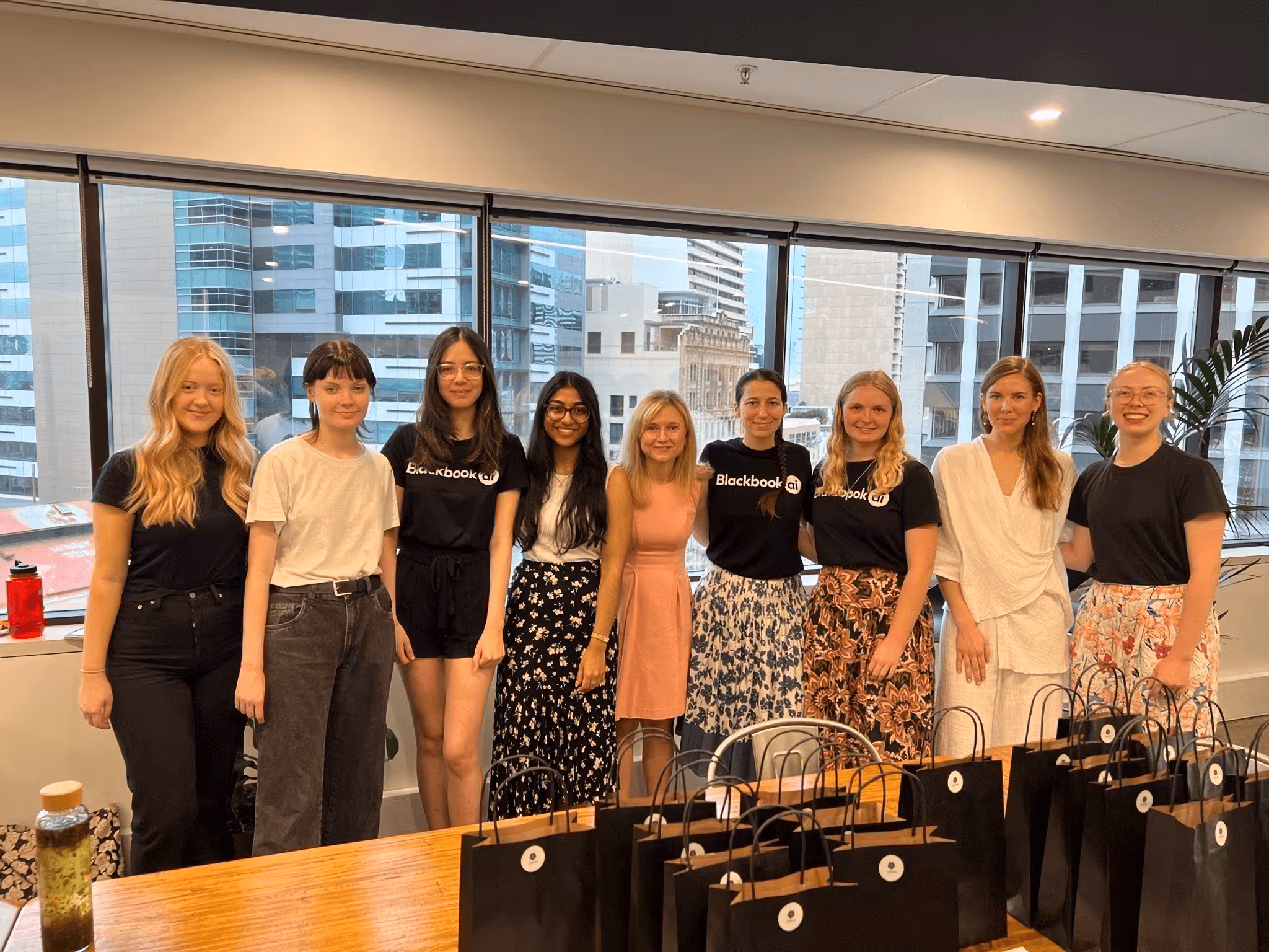Group of nine women standing indoors in front of large windows with city buildings outside, smiling behind a wooden table with black gift bags.