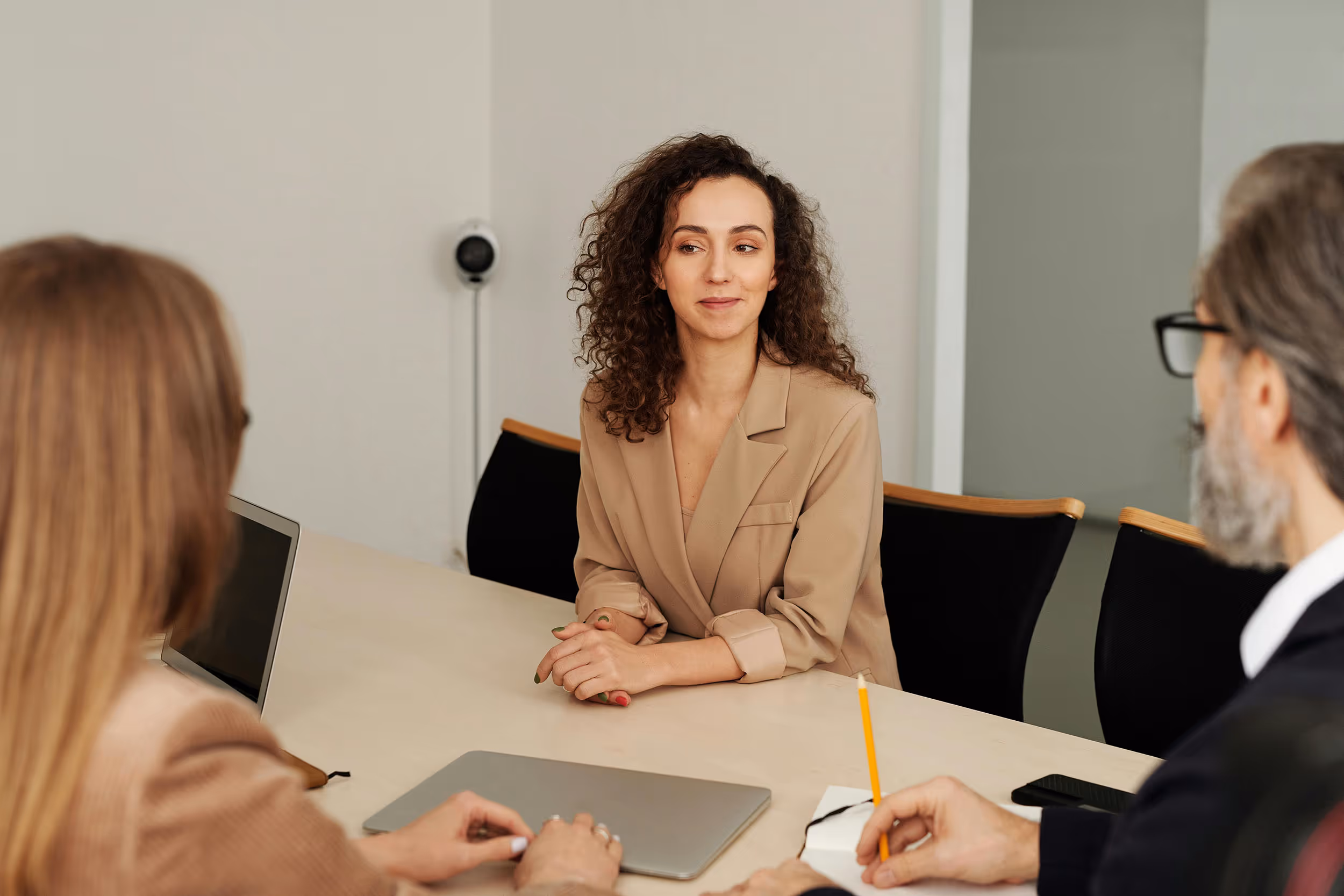 Young woman in a beige blazer sitting at a table speaking with two colleagues in a meeting room.