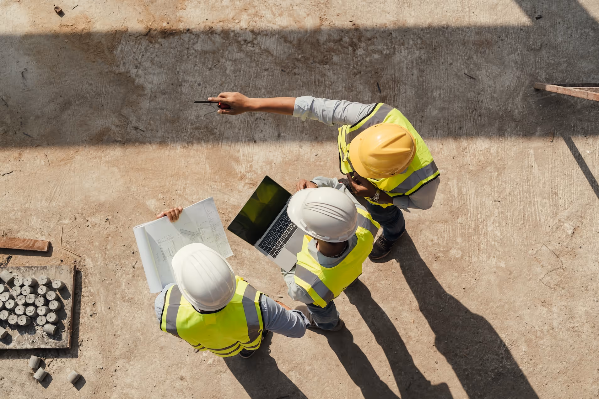 Three construction workers wearing safety helmets and reflective vests reviewing blueprints and a laptop on a concrete floor.