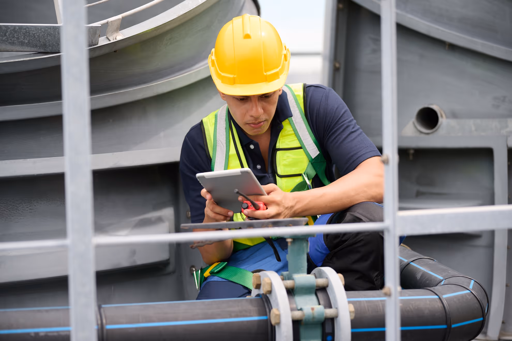 Worker wearing a yellow hard hat and safety vest inspecting equipment and using a tablet on an industrial site.
