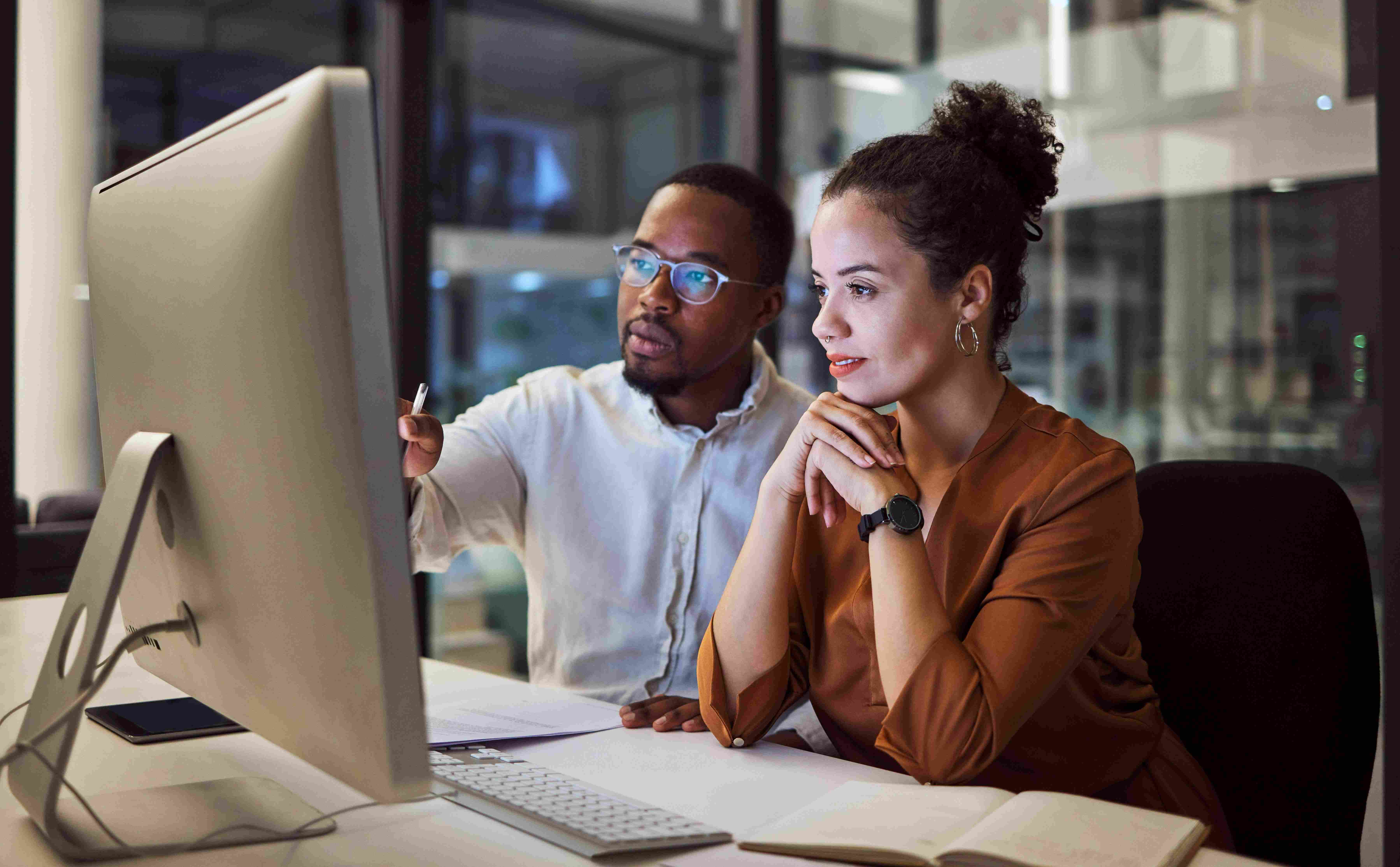 Two colleagues working together at a computer in an office, one pointing at the screen and the other attentively watching.