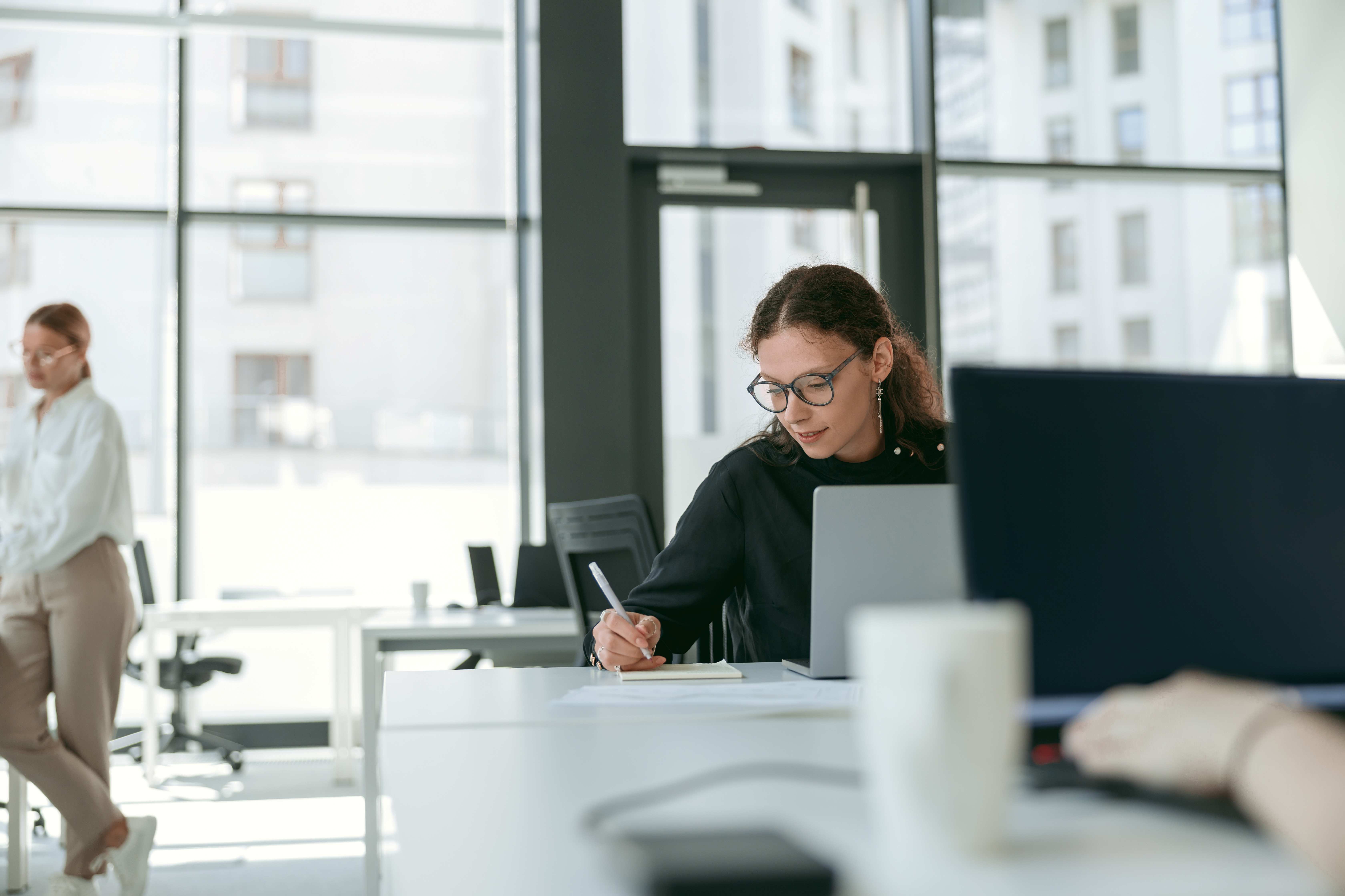 Woman with glasses writing on a notepad while sitting at a desk with a laptop in a bright modern office.
