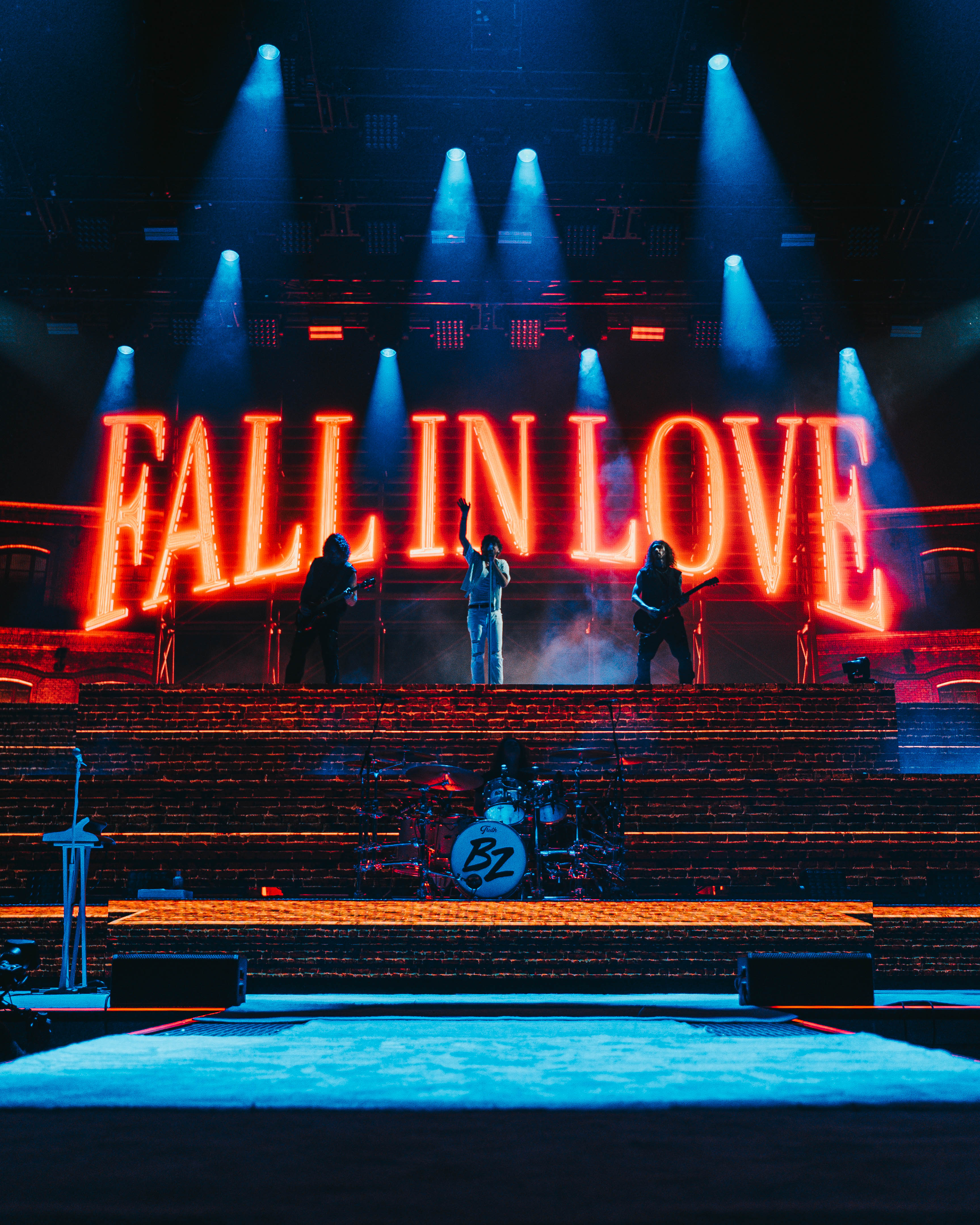 A stage with three man on a platform with the words FALL IN LOVE in neon behind them