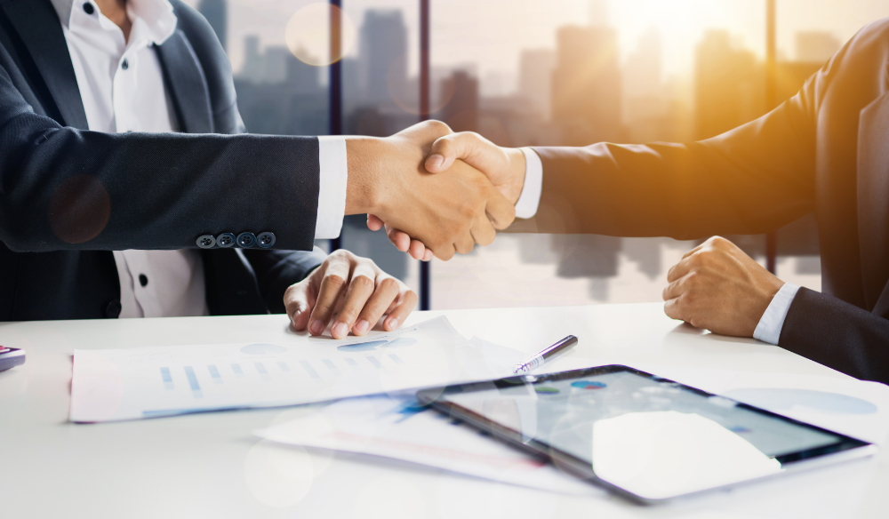 handshaking businessmen with documents and a tablet on top of the table