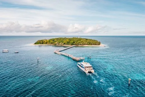 Boat approaching a dock connected to a small green island surrounded by blue ocean under a partly cloudy sky.