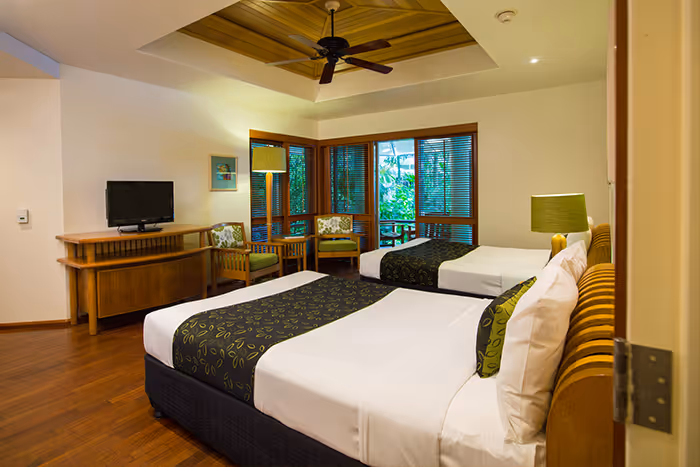Hotel room with two beds, a wooden TV stand, green-accented chairs near a corner window, and a ceiling fan.