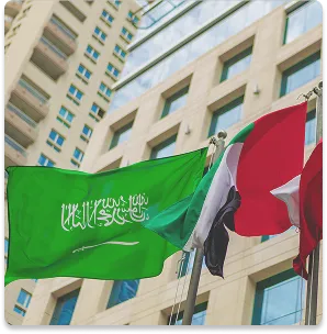Saudi Arabian and other flags waving in front of a modern office building with many windows.