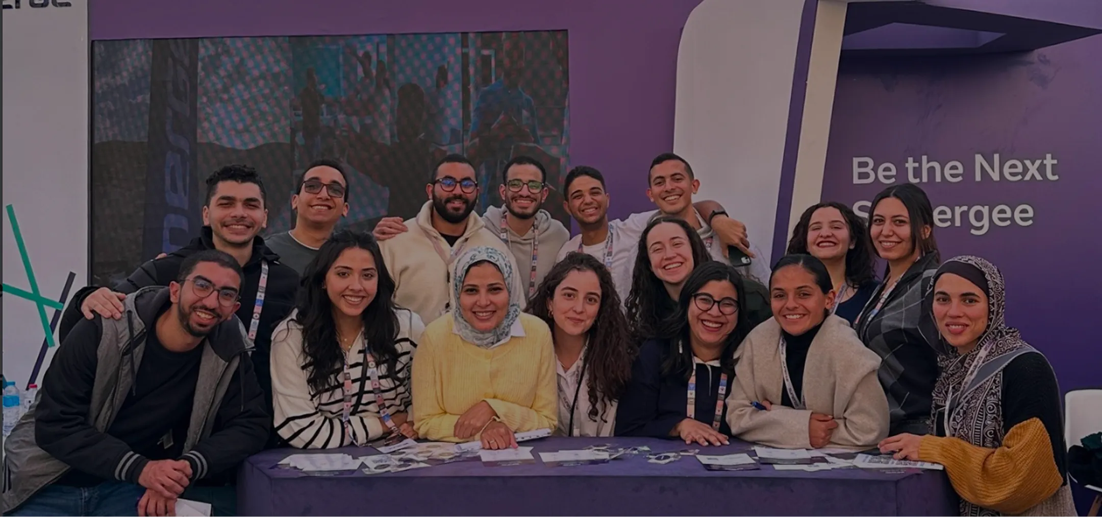 Group of smiling young people leaning on a table with a purple background that reads Be the Next Sumergee.