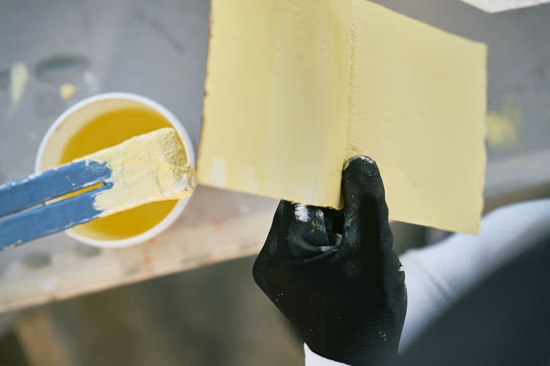 Hand in schwarzem Handschuh hält ein Stück Holz, das mit gelber Farbe gestrichen wird.